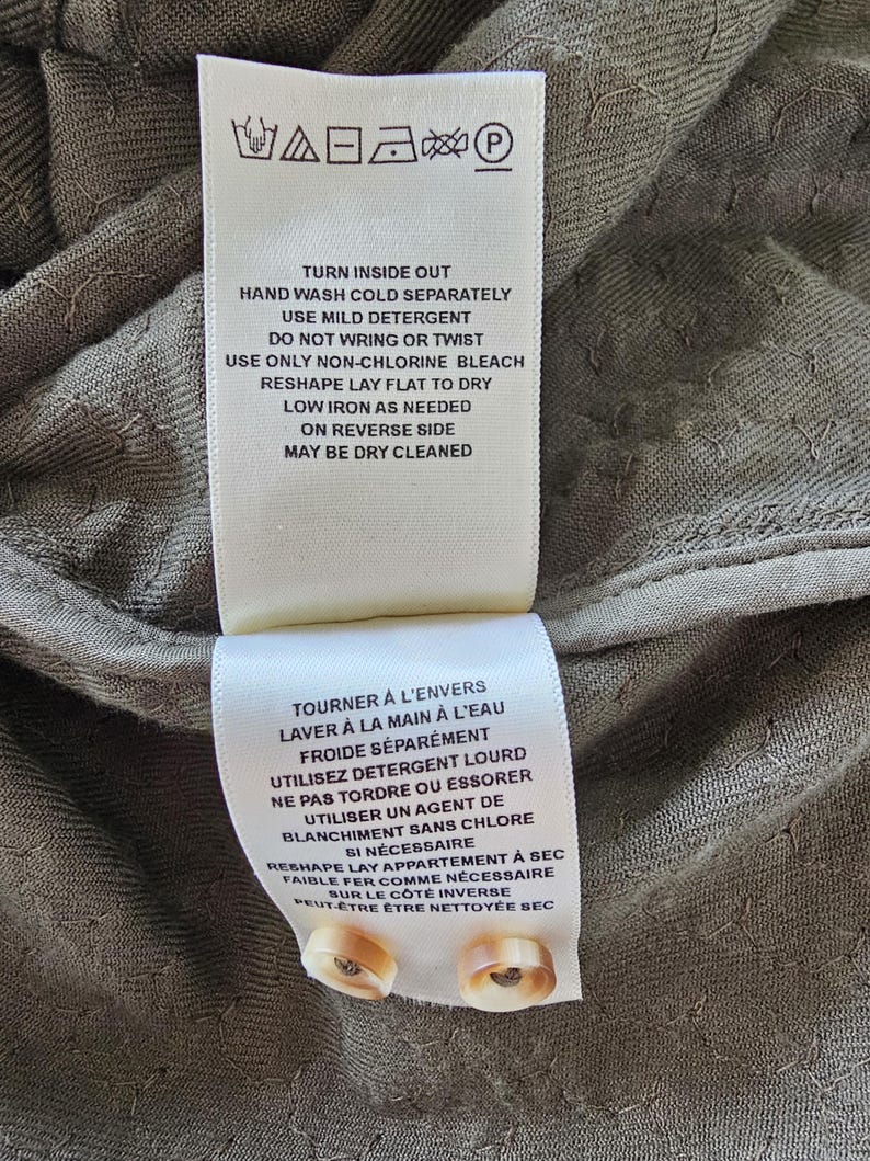 May include: Close-up of two white fabric tags with care instructions in English and French. The tags are attached to a textured, olive-green fabric. The English tag includes washing and ironing instructions. Two beige buttons are visible.