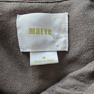 May include: Close-up of a garment tag with the brand name "Maeve" in gold lettering. The tag is white and rectangular, with the size "M" and "Made in India" printed below. The fabric is a textured, olive-green color.