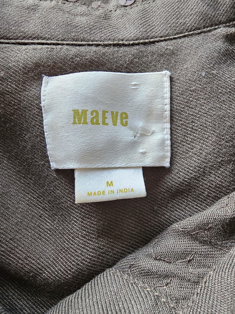 May include: Close-up of a garment tag with the brand name "Maeve" in gold lettering. The tag is white and rectangular, with the size "M" and "Made in India" printed below. The fabric is a textured, olive-green color.