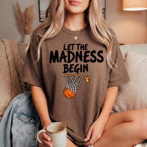 May include: Brown t-shirt with the text "LET THE MADNESS BEGIN" in black. A basketball net with a basketball is printed below the text. The shirt is a casual style.