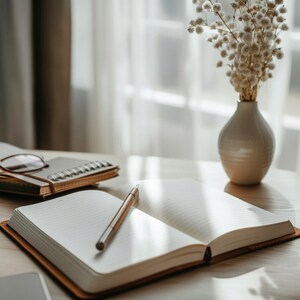 May include: An open notebook with lined pages and a pen lies on a wooden surface. A small vase with dried flowers and a closed notebook with glasses are in the background. The scene is illuminated by soft, natural light, creating a calm atmosphere.