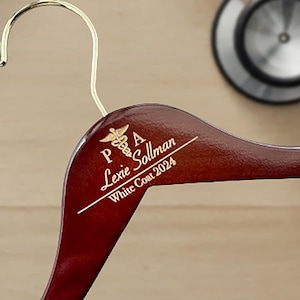 May include: A dark red wooden hanger with a gold inscription that reads "PA Lexie Sollman White Coat 2024". The inscription also includes a small gold medical symbol.