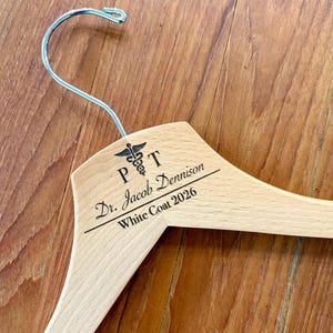 May include: A wooden clothes hanger with a silver hook. The hanger is engraved with the letters "PT", a medical symbol, and the text "Dr. Jacob Dennison White Coat 2026". The hanger is made of light-colored wood.