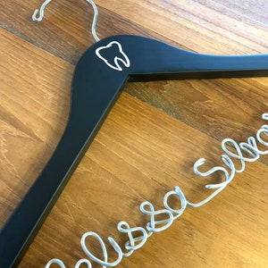May include: Black wooden hanger with a white tooth graphic and a silver wire name "Melissa" hanging from it.