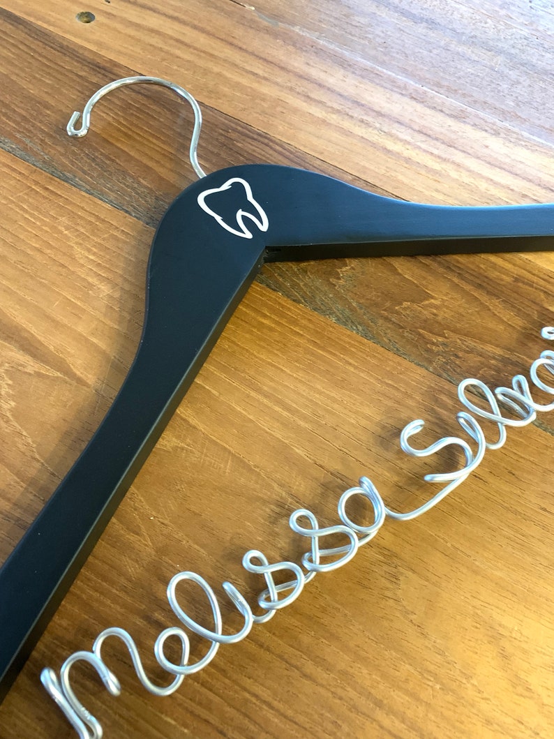 May include: Black wooden hanger with a white tooth graphic and a silver wire name "Melissa" hanging from it.