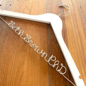 Può includere: Un appendiabiti in legno bianco con il nome "Beth Brown" scritto in filo d'argento.