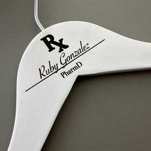 May include: White wooden hanger with a black inscription that reads "Rx Ruby Gonzalez PharmD".