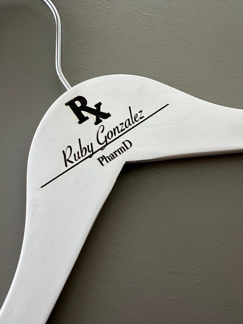 May include: White wooden hanger with a black inscription that reads "Rx Ruby Gonzalez PharmD".