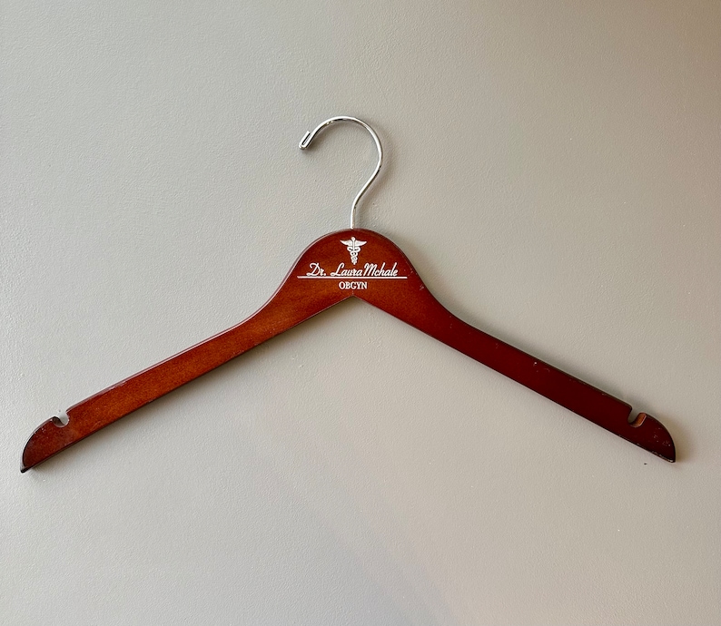 May include: A brown wooden hanger with a silver hook and a white inscription that reads "Dr. Laura Michale OB/GYN" with a medical symbol.