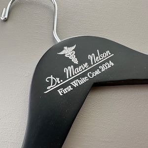 May include: Black wooden hanger with a silver medical symbol and the text "Dr. Maeve Nelson First White Coat 2024" engraved on it.
