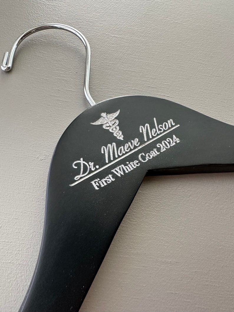 May include: Black wooden hanger with a silver medical symbol and the text "Dr. Maeve Nelson First White Coat 2024" engraved on it.