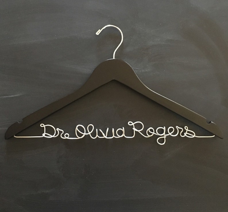 May include: Black wooden hanger with a silver wire inscription that reads "Dr. Olivia Rogers".