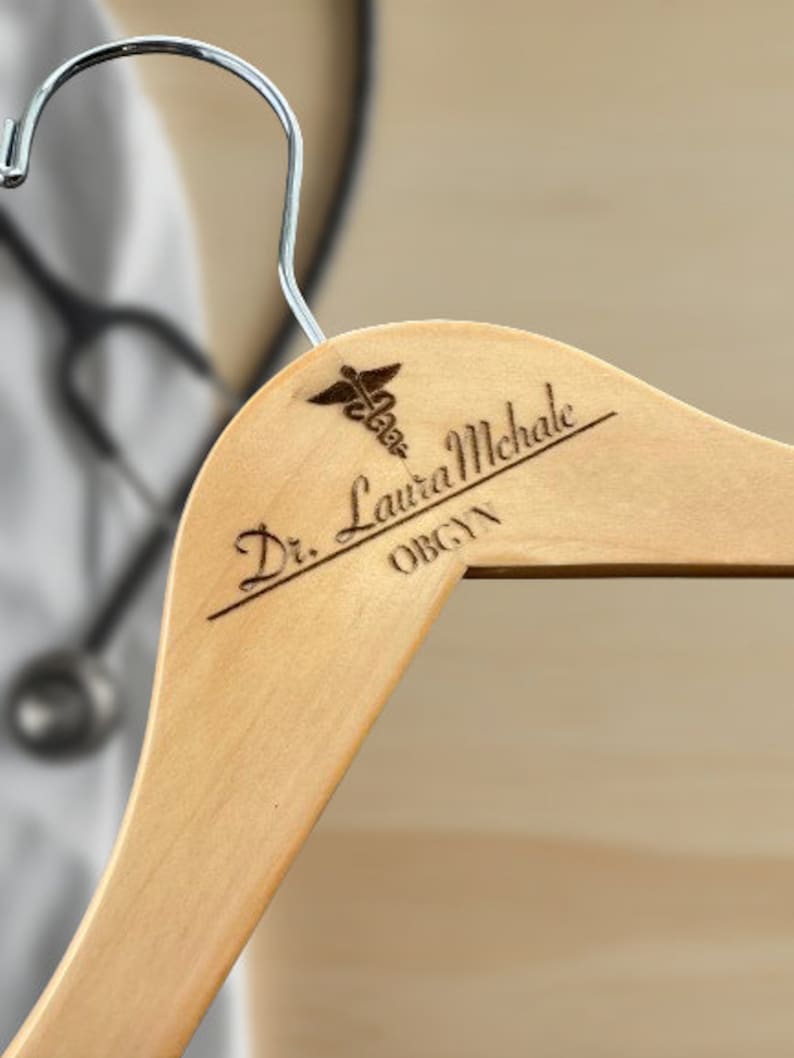 May include: A wooden hanger with a personalized inscription that reads "Dr. Laura Michale OBGYN" and a medical symbol.