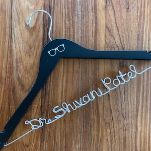 May include: A black wooden hanger with a silver wire inscription that reads "Dr. Shuvam Patel". The hanger has a small pair of glasses icon printed on the top.