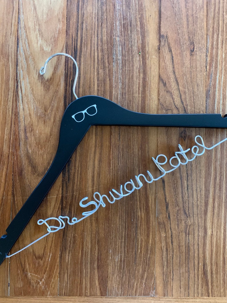 May include: A black wooden hanger with a silver wire inscription that reads "Dr. Shuvam Patel". The hanger has a small pair of glasses icon printed on the top.