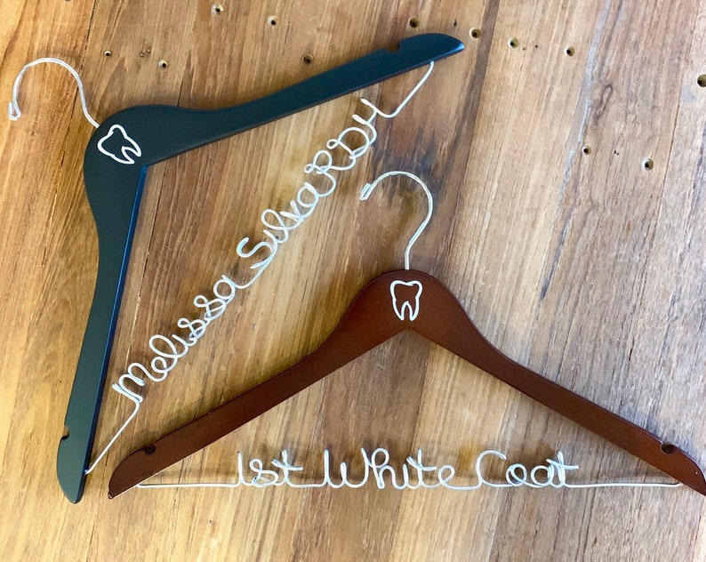 May include: Two wooden hangers with a tooth design on the top. One hanger is black and says "Melissa Sullivan" in white wire. The other hanger is brown and says "1st White Coat" in white wire.