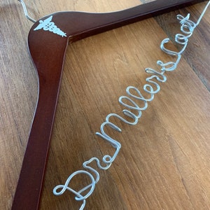 May include: A brown wooden hanger with a silver wire inscription that reads "Dr. Miller's Coat". The hanger has a white medical symbol on the top.