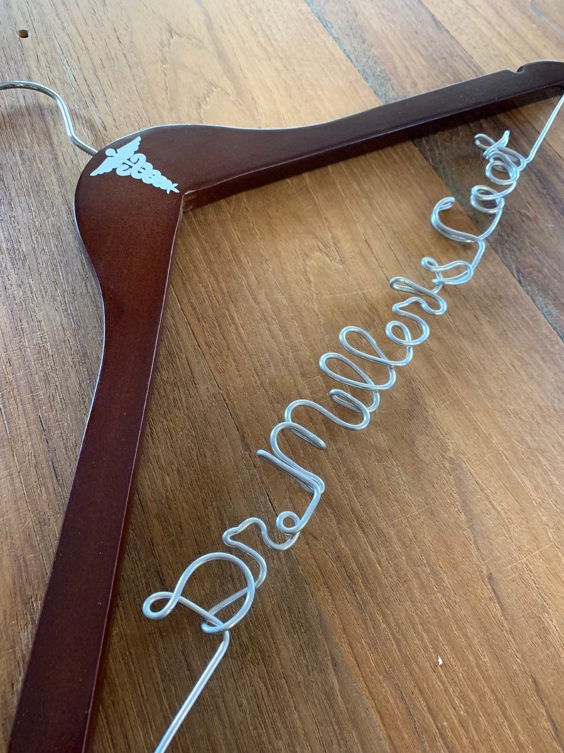 May include: A brown wooden hanger with a silver wire inscription that reads "Dr. Miller's Coat". The hanger has a white medical symbol on the top.