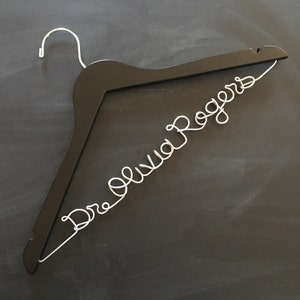May include: A black wooden hanger with the name "Dr. Olivia Rogers" written in silver wire.