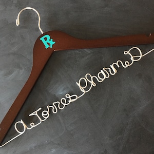 May include: A brown wooden hanger with a turquoise "R" on the top and the words "Dr. Torres Pharmacy" written in silver wire.