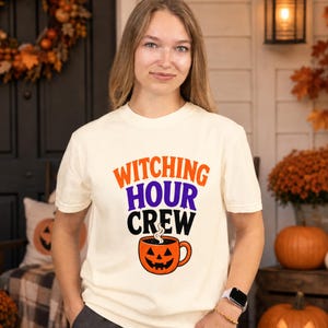 May include: A cream-colored short-sleeve t-shirt featuring the text "WITCHING HOUR CREW" in orange, purple, and black, with a graphic of a smiling jack-o'-lantern coffee mug below the text, showing steam rising from the top of the mug.