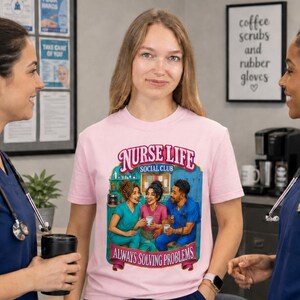 May include: A light pink t-shirt featuring a graphic illustration of three people in scrubs sitting at a table with coffee mugs. The text reads Nurse Life Social Club Always Solving Problems. The shirt is worn by a person in a medical office setting.