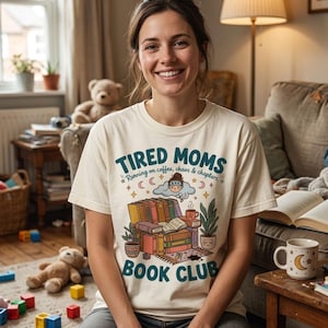 Op de afbeelding: Een vrouw draagt een crèmekleurig t-shirt met de tekst "TIRED MOMS BOOK CLUB" in teal en oranje. Het shirt heeft een afbeelding van boeken, een koffiekopje en een wolk met een persoon erin. De vrouw lacht.