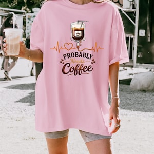 May include: A light pink t-shirt featuring a graphic of an IV bag filled with coffee, a heart-shaped EKG line, and the text "PROBABLY Needs Coffee" in brown and orange. The design includes small coffee bean illustrations surrounding the text.