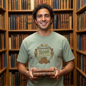 Pode incluir: Uma pessoa vestindo uma camiseta verde com o texto "Treasure Hunter Book Division" segurando dois livros antigos. O fundo apresenta uma biblioteca com muitos livros antigos em prateleiras de madeira.