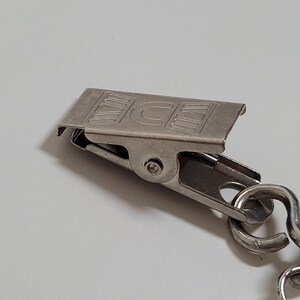 May include: A close-up of a silver-colored metal clip with a chain. The clip has the letters "ICE" printed on it. The clip is attached to a chain with a curved design.
