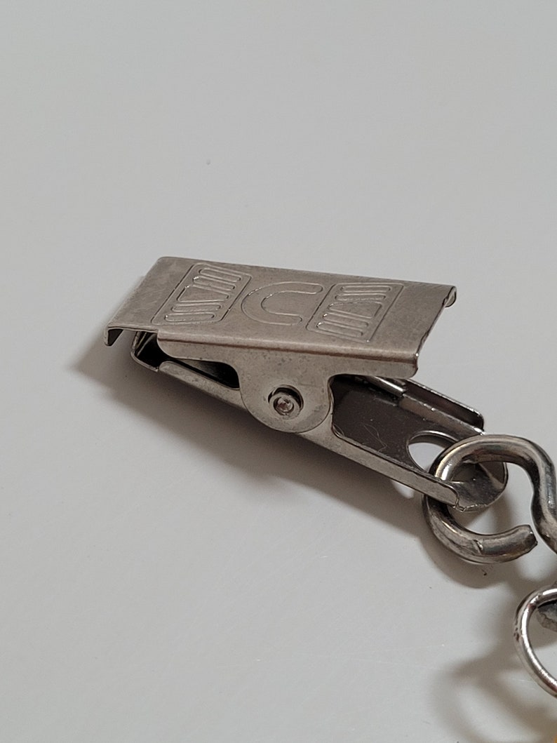 May include: A close-up of a silver-colored metal clip with a chain. The clip has the letters "ICE" printed on it. The clip is attached to a chain with a curved design.