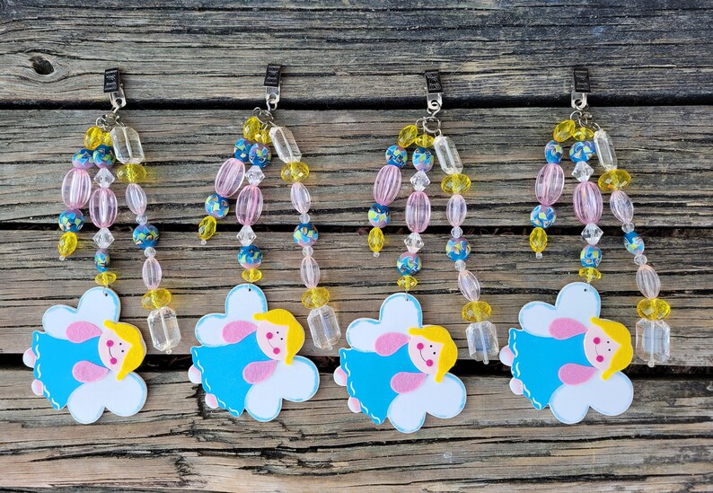 May include: Four decorative beaded angel ornaments. Each ornament features a cloud-shaped design with a smiling angel in a blue dress, yellow hair, and pink cheeks. The beaded strands include pink, yellow, and blue beads, and clear rectangular beads.
