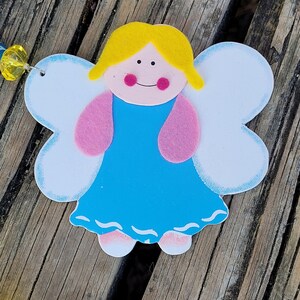 May include: A wooden angel decoration with a light blue dress, pink arms, and white wings. The angel has yellow hair, pink cheeks, and a smiling face. The angel is on a wooden surface.