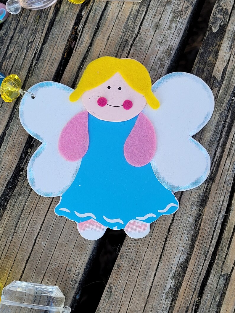 May include: A wooden angel decoration with a light blue dress, pink arms, and white wings. The angel has yellow hair, pink cheeks, and a smiling face. The angel is on a wooden surface.