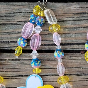 May include: A beaded ornament with a variety of colorful beads. The beads include yellow, pink, clear, and blue with a floral design. The ornament is hanging from a silver hook. The background is a weathered wooden surface.