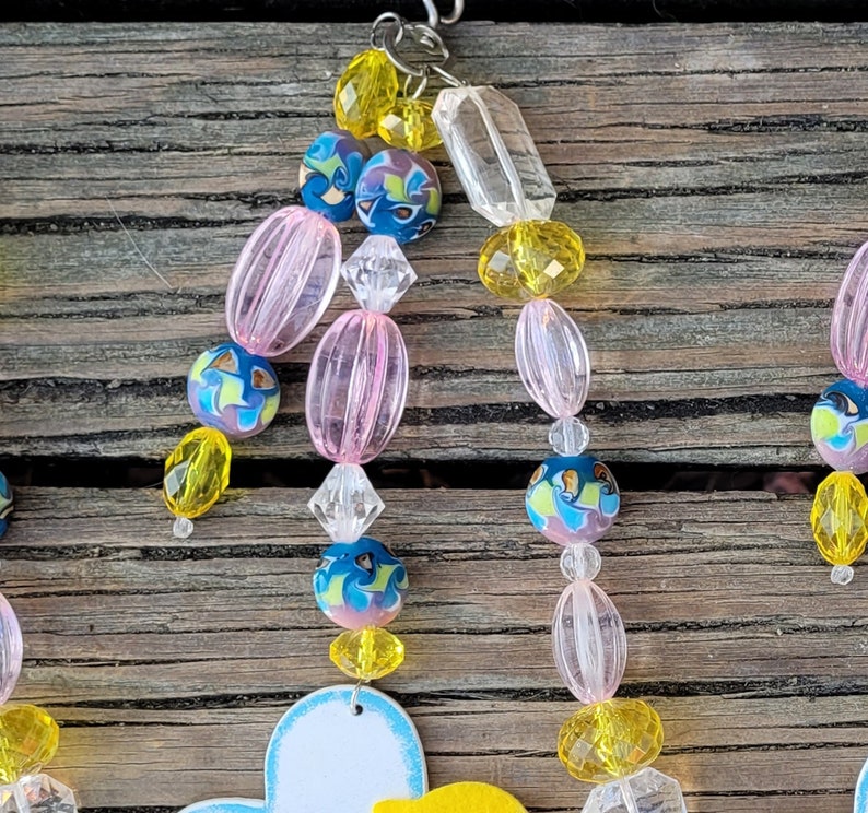 May include: A beaded ornament with a variety of colorful beads. The beads include yellow, pink, clear, and blue with a floral design. The ornament is hanging from a silver hook. The background is a weathered wooden surface.