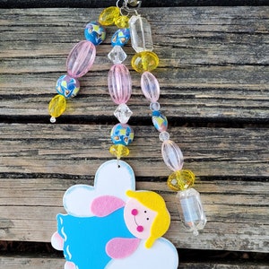 May include: A decorative angel ornament with a blue dress, pink arms, and yellow hair. The angel is attached to a beaded strand with pink, yellow, and blue beads, and a metal clip at the top. The ornament is designed to hang.
