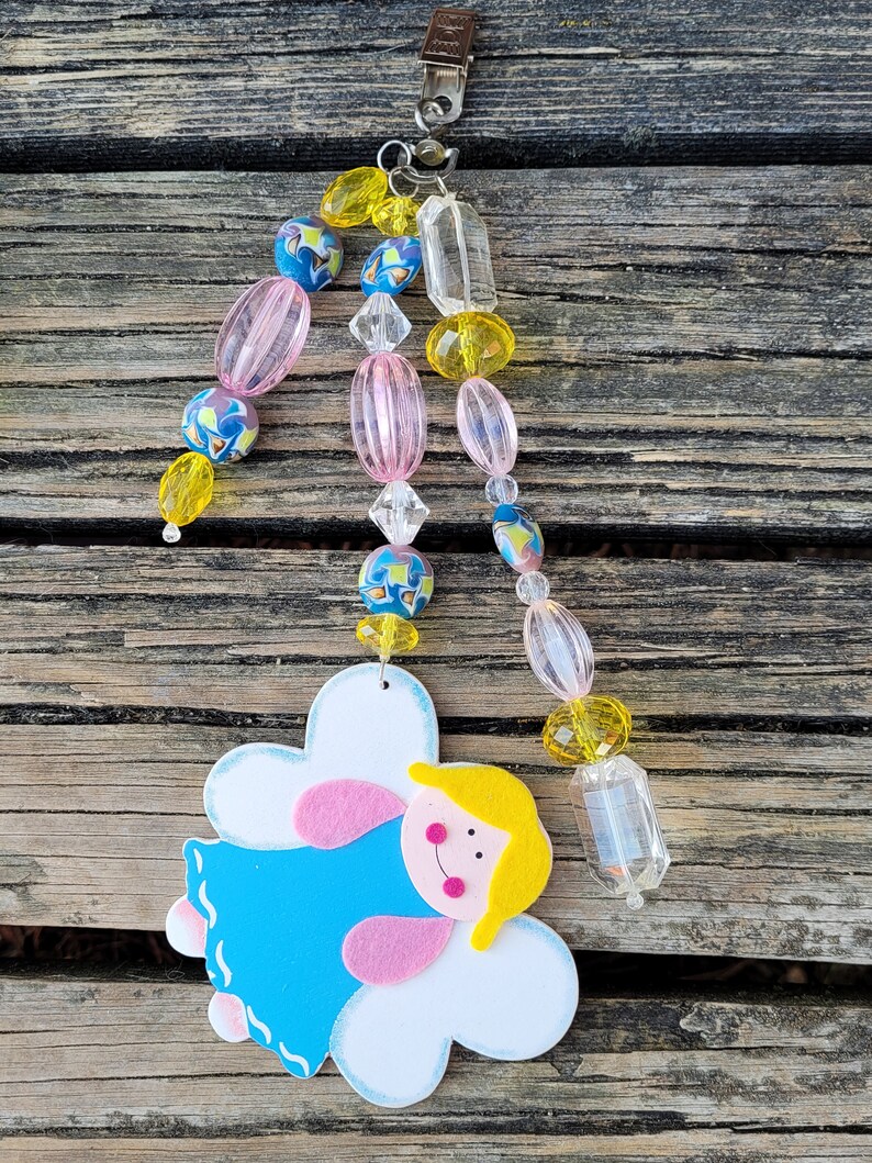 May include: A decorative angel ornament with a blue dress, pink arms, and yellow hair. The angel is attached to a beaded strand with pink, yellow, and blue beads, and a metal clip at the top. The ornament is designed to hang.