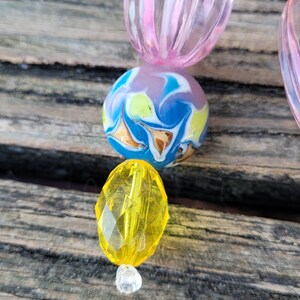 May include: Close-up of a beaded accessory featuring a mix of colorful beads. The design includes a pink, ribbed oval bead, a marbled blue, yellow, and white round bead, and a faceted yellow bead. A clear, small bead is at the bottom.