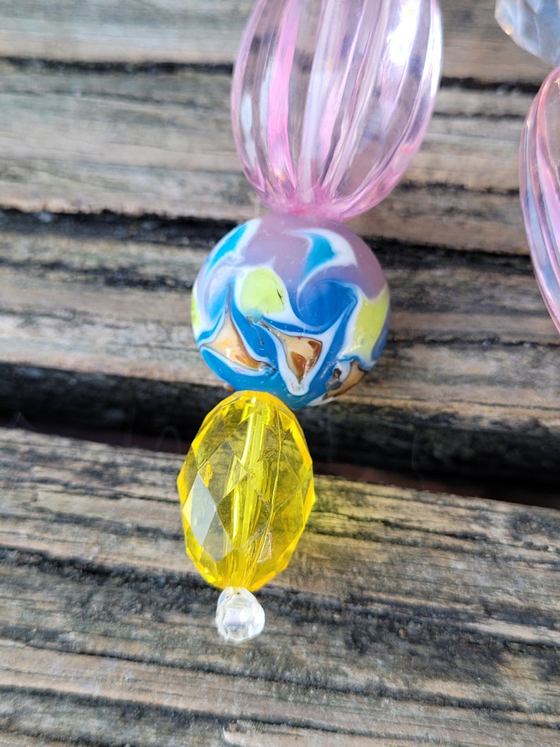 May include: Close-up of a beaded accessory featuring a mix of colorful beads. The design includes a pink, ribbed oval bead, a marbled blue, yellow, and white round bead, and a faceted yellow bead. A clear, small bead is at the bottom.