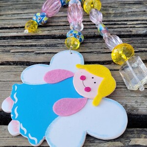 May include: A handmade angel-shaped ornament with a blue dress, pink accents, and a yellow head. The ornament is attached to a beaded strand with pink, yellow, and blue beads. A clear, rectangular charm hangs from the strand.