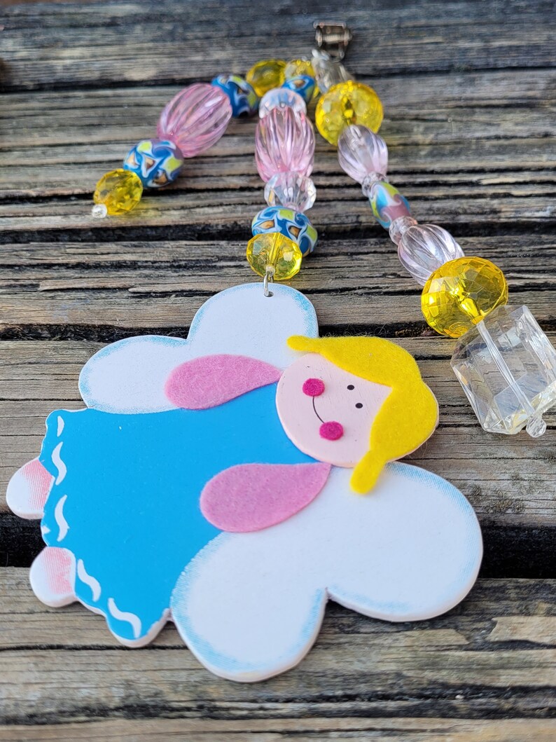 May include: A handmade angel-shaped ornament with a blue dress, pink accents, and a yellow head. The ornament is attached to a beaded strand with pink, yellow, and blue beads. A clear, rectangular charm hangs from the strand.