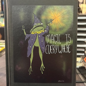 Peut inclure: Une couverture de livre noire avec une illustration d'une grenouille verte habillée en magicien, tenant une baguette avec une étoile. Le texte "MAGIC IS EVERYWHERE" est écrit en blanc. La grenouille porte une robe et un chapeau violets.