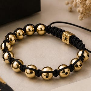 Op de afbeelding: Een gouden kralenarmband met zwart koord, gepresenteerd met een zwarte geschenkdoos en een kaart met de tekst "Especially for You". De armband heeft gouden kralen en een decoratieve vierkante kraal.
