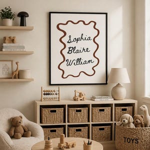 May include: A framed print with the names Sophia, Blaire, and William in black script, outlined by a brown wavy shape. The room features a neutral color palette with wooden shelves, a lamp, and a storage unit with woven baskets. Toys and decorative items are also visible.