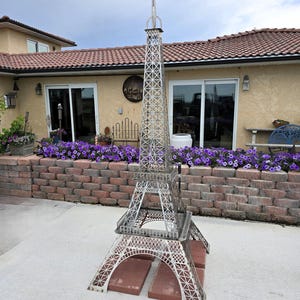 May include: A detailed metal Eiffel Tower sculpture, silver in color, stands on a base of red bricks. The tower's intricate lattice design is visible, with a tall spire reaching towards the sky. The sculpture is an outdoor decorative item.