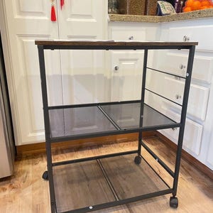 Puede incluir: Un carrito de cocina rodante de metal negro con una tapa de madera marrón oscuro y estantes de malla. El carrito tiene cuatro ruedas para facilitar el movimiento y está diseñado para el almacenamiento y la organización. El carrito está en una cocina.