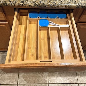 May include: A wooden drawer organizer with multiple compartments. The organizer is made of light-colored bamboo and features sections for utensils and containers. Blue-lidded containers and a white spoon are visible. The drawer is set in a wooden cabinet with a granite countertop.
