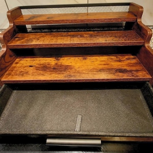 May include: A wooden desk organiser with three shelves and a pull-out drawer. The shelves are made of stained wood, and the top shelf has a black wire mesh. The drawer is lined with a dark grey fabric.