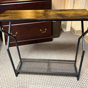 Könnte beinhalten: Rechteckiger Beistelltisch mit dunkler Holzoptik-Oberfläche und schwarzem Metallrahmen. Der Tisch hat ein unteres Regal und ein einzigartiges, abgewinkeltes Beindesign. Die Oberseite ist etwa 100 cm lang.